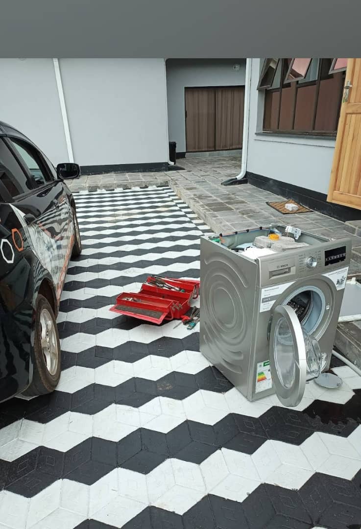 Front-loading washing machine opened on a chequered driveway beside a red toolbox